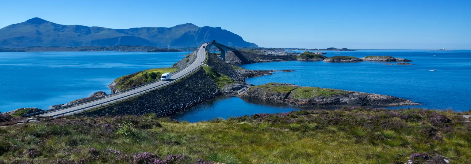 Ein Sunny Cars Mietwagen fährt auf einer Straße über eine Brücke über dem Meer mit Bergen im Hintergrund 