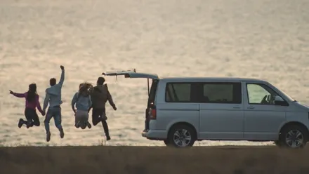 Freundesgruppe springt freudig am Strand in die Luft, 7-Sitzer parkt daneben