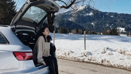Person sitzt auf der geöffneten Kofferraumkante eines Autos am Straßenrand vor verschneiter Landschaft mit Bergen im Hintergrund