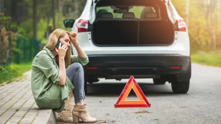 Auto mit Panne kurz vor dem Urlaub – Person telefoniert am Straßenrand neben geöffnetem Kofferraum und Warndreieck