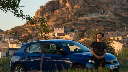 Person lehnt an einem Mietwagen vor einer Berglandschaft und blickt nach oben in den Himmel