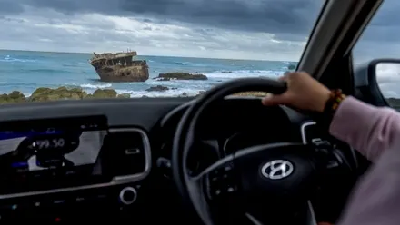 Ein Mann in einem Auto mit rechts montiertem Lenkrad blickt an der Küste auf ein Schiffswrack