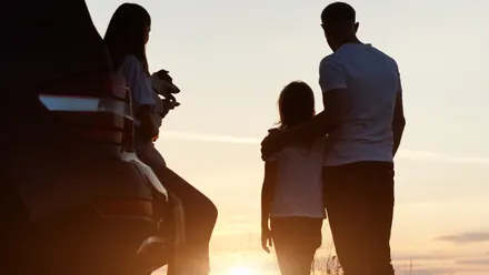 Drei Personen stehen neben einem geöffneten Auto bei Sonnenuntergang, eine Person hält einen kleinen Hund im Arm