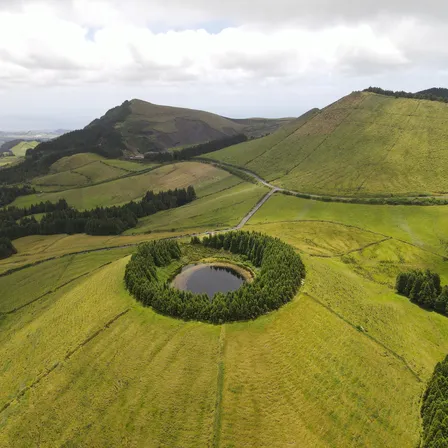 Luftaufnahme einer grünen Hügellandschaft auf Terceira mit einem ringförmig von Bäumen umgebenen kleinen See in der Mitte