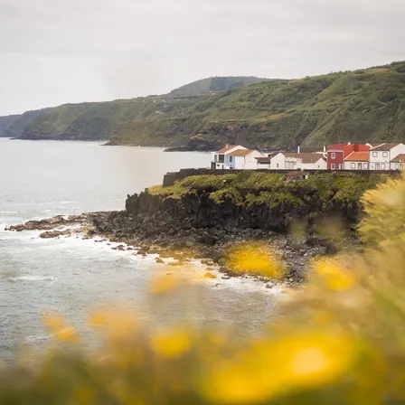 Kleine Häuser auf einer Klippe an der Küste von São Miguel, umgeben von grünen Hügeln und schwarzer Lavaküste, mit Meer im Vordergrund