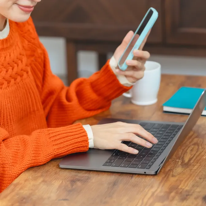 Frau sitzt an einem Laptop und hält ein Smartphone in der Hand