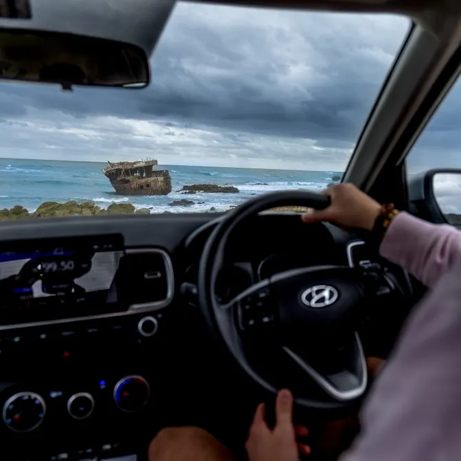 Ein Mann in einem Auto mit rechts montiertem Lenkrad blickt an der Küste auf ein Schiffswrack