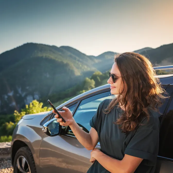 Woman looking at her smartphone, with a parked rental car in the mountains in the background